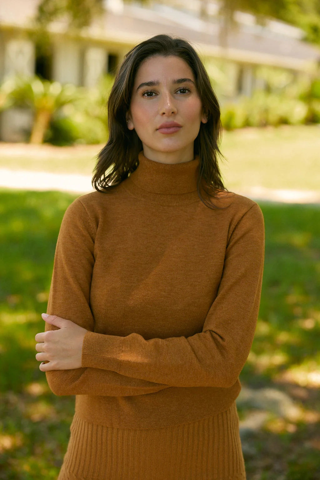 A woman wearing a warm rust-brown ribbed turtleneck sweater stands outdoors with her arms crossed, set against a softly blurred green lawn and house background.