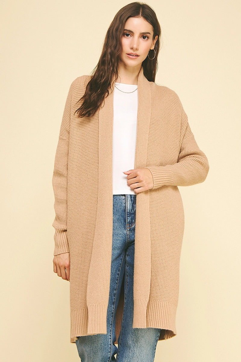 Woman wearing a long beige cardigan over a white top and blue jeans on a beige background