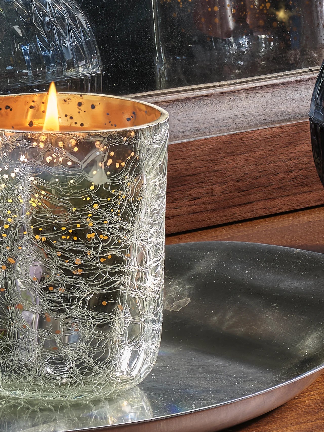 Candle in a textured glass holder on a reflective surface with a wooden background
