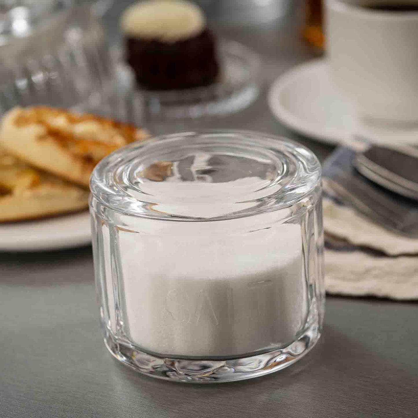 Clear glass container with white powder on a table with food and coffee in the background