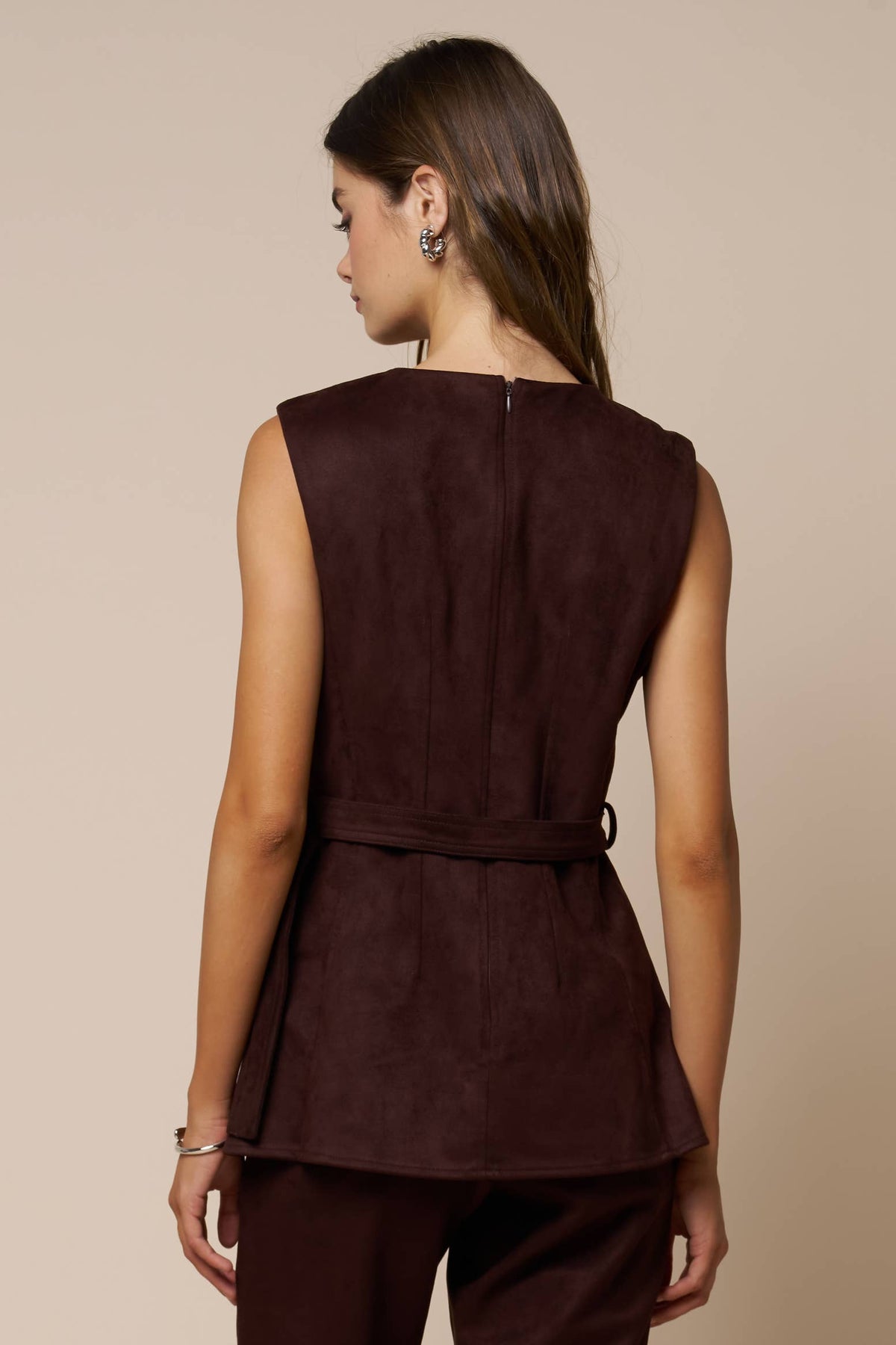 A woman shown from the back wearing a sleeveless dark plum-brown peplum top with a matching tie belt, against a plain beige studio background.