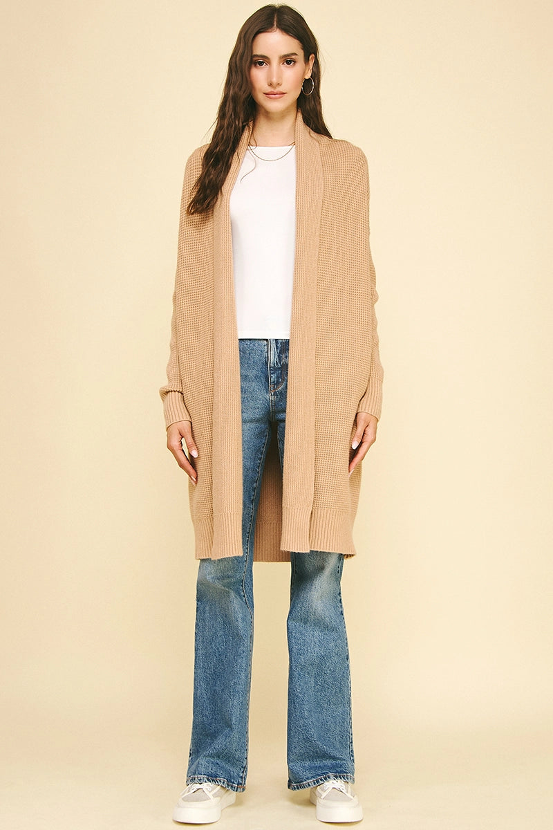 Woman wearing a beige cardigan, white shirt, blue jeans, and white sneakers on a beige background