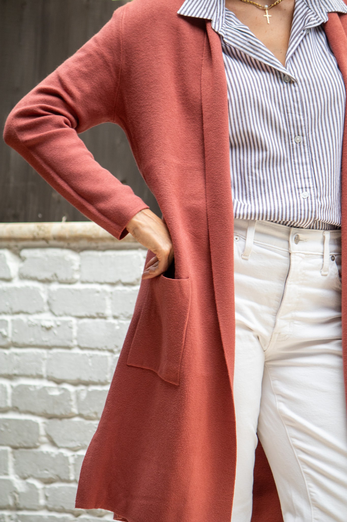 Person wearing a long rust-colored cardigan over a striped shirt with white pants against a brick wall.