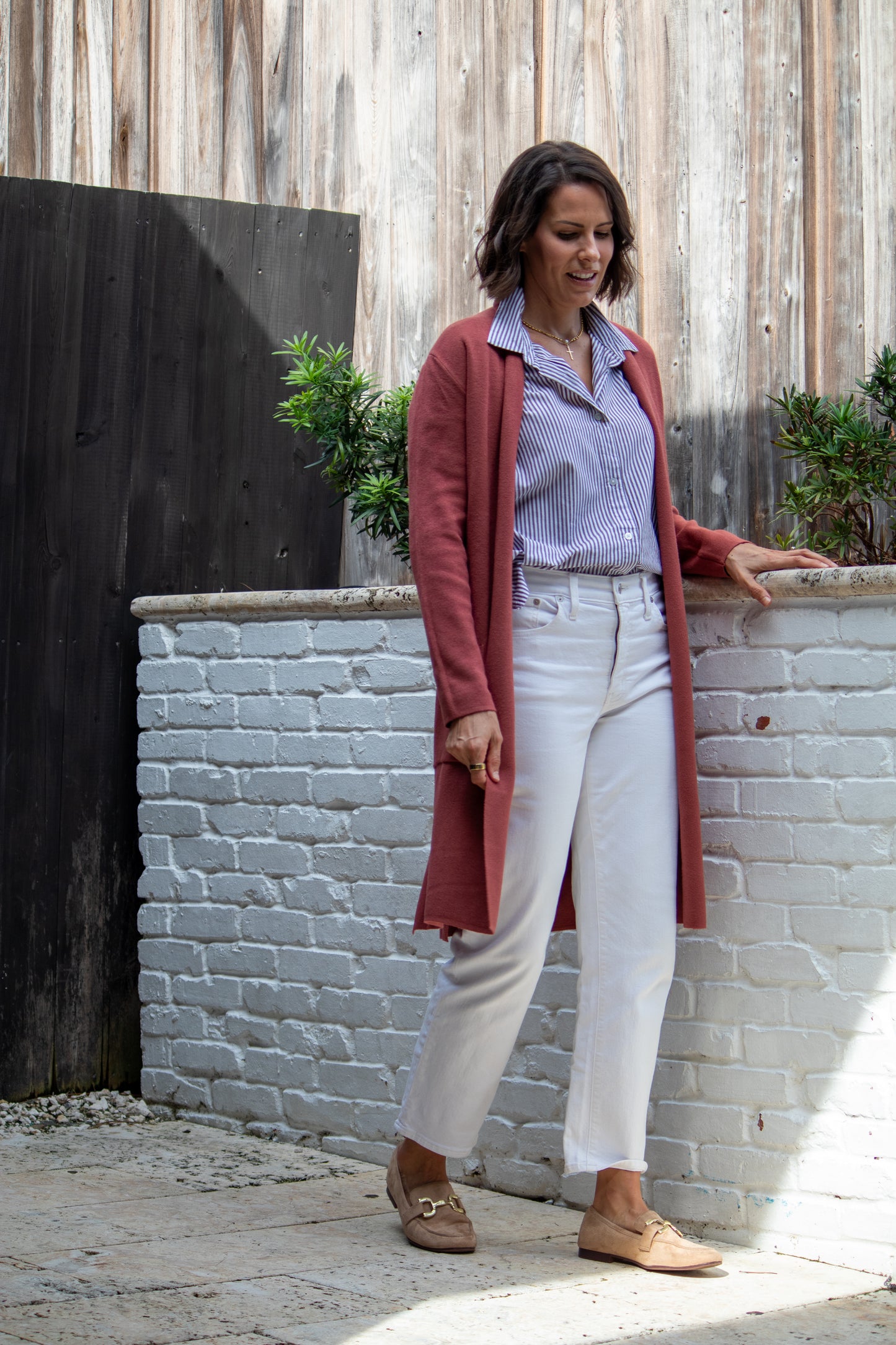 Woman standing against a white brick wall wearing a long red cardigan, light blue shirt, white pants, and brown shoes.