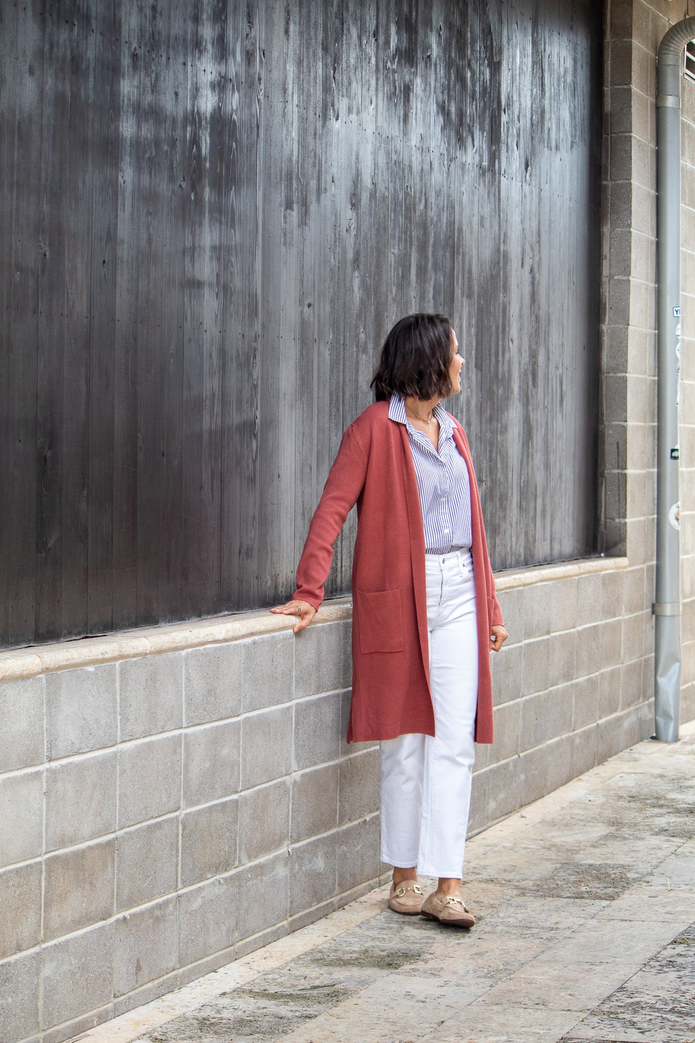 Woman standing against a textured wall wearing a red coat and white pants.