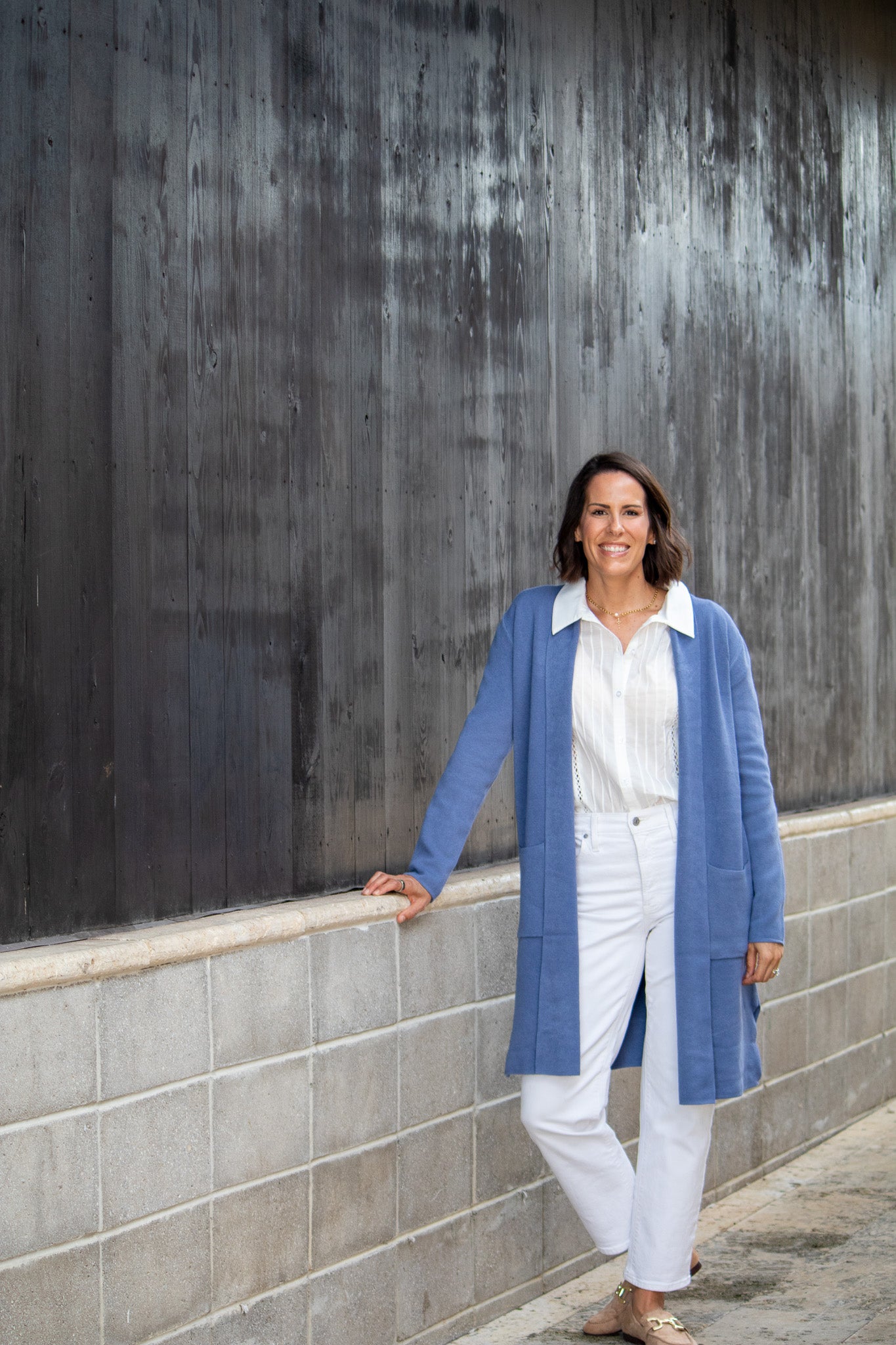 Woman in a blue cardigan and white pants standing against a textured wall.