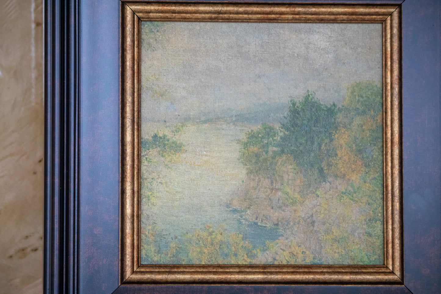 Framed painting of a landscape with trees and water on a blue wall.