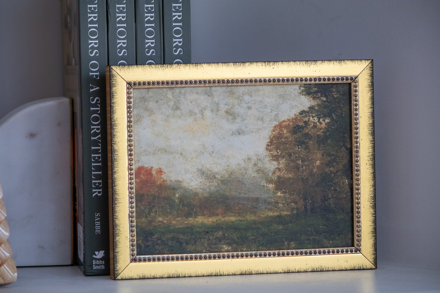 Framed landscape painting on a shelf with books in the background
