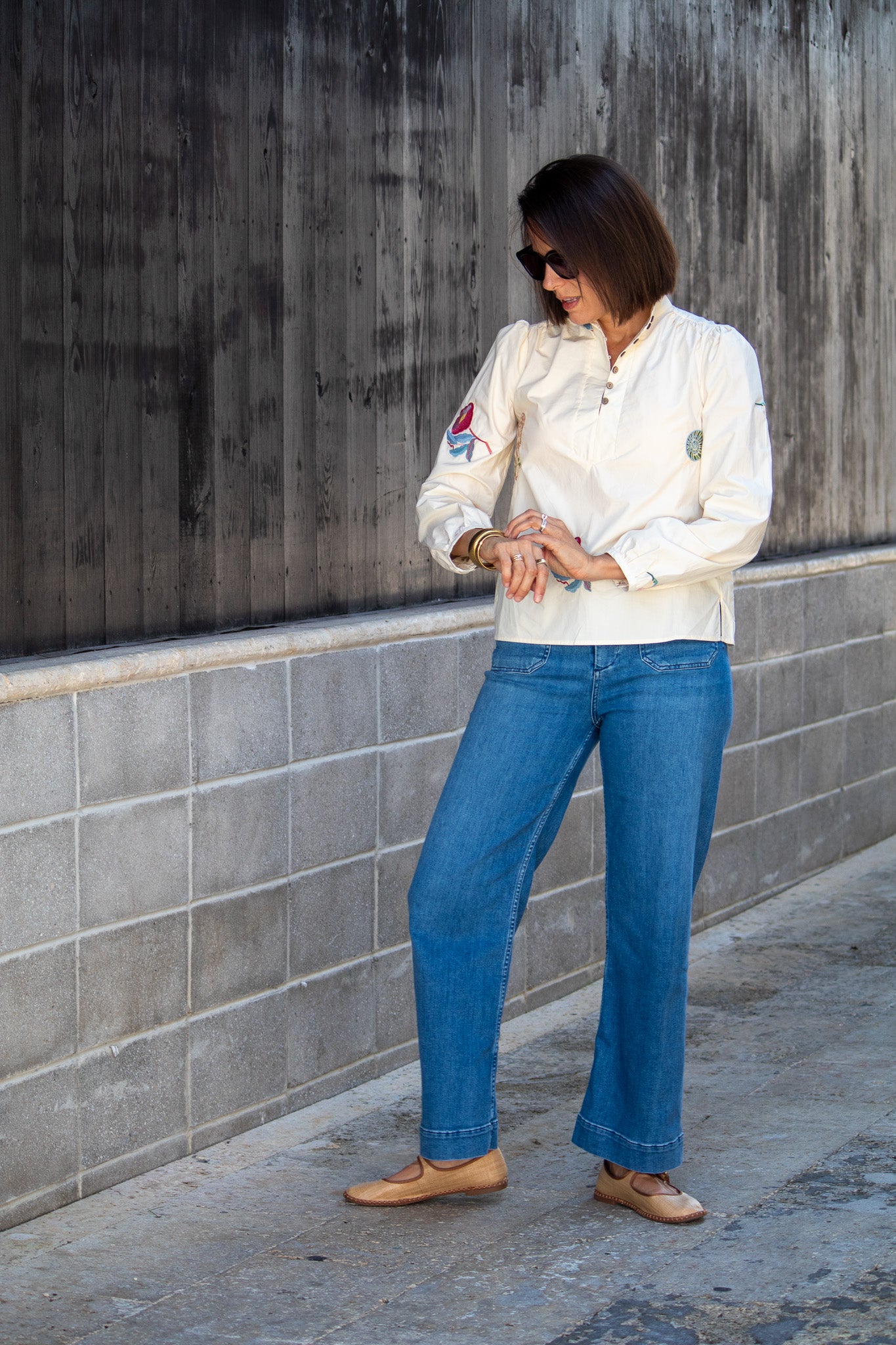 Woman wearing a white top and blue jeans standing against a gray wall.