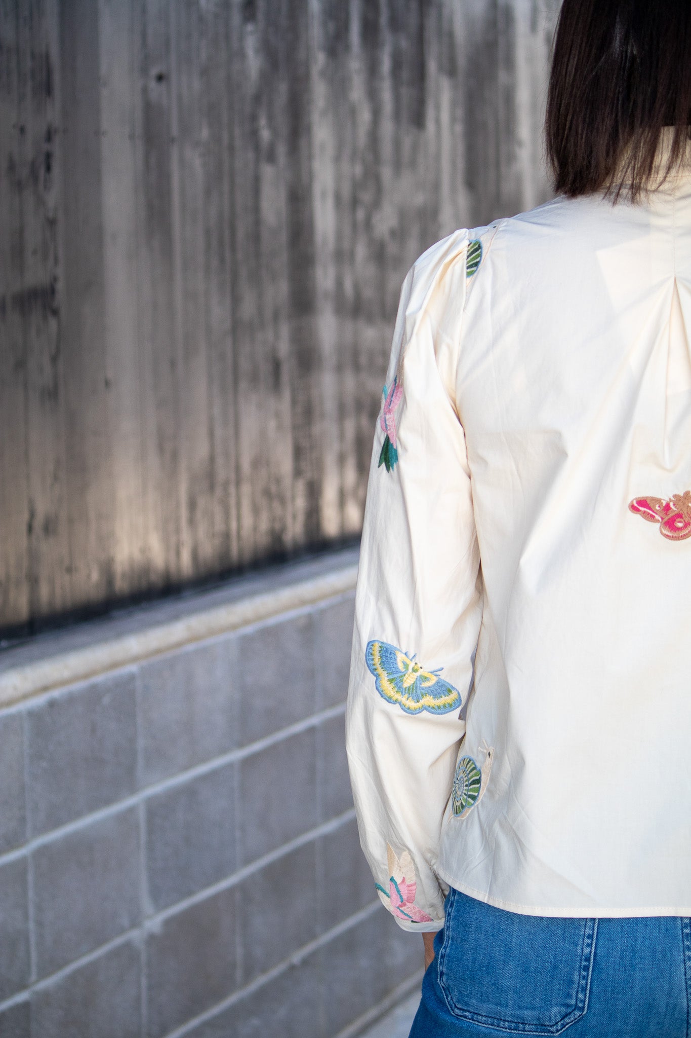 Person wearing a white embroidered blouse with blue jeans against a concrete wall.