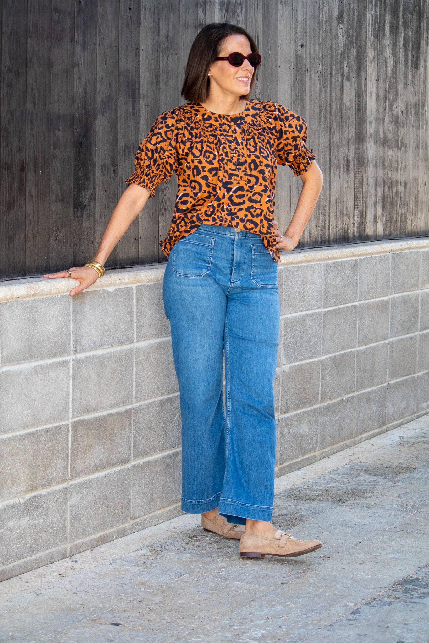 Woman wearing a leopard print top and blue jeans leaning against a wall.