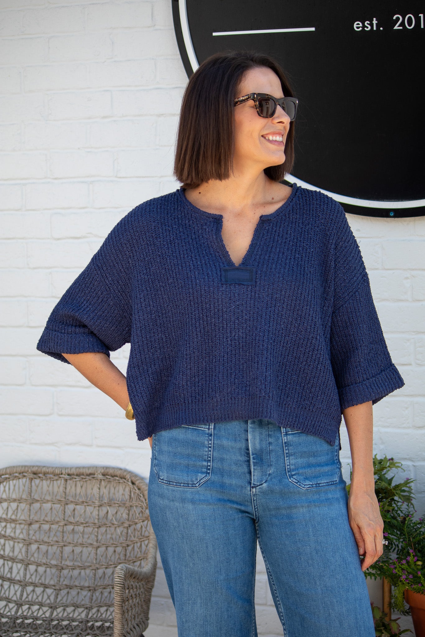 Woman wearing a blue textured top and jeans standing against a white brick wall.