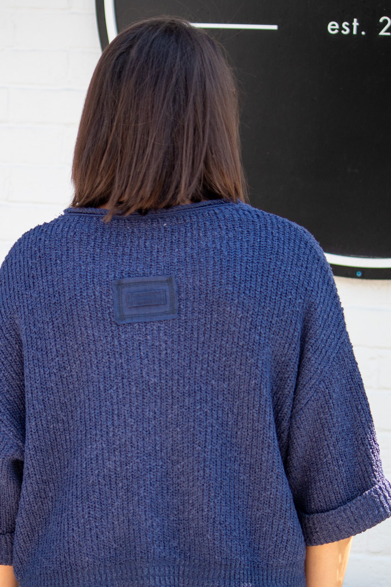 Person wearing a blue textured sweater with a logo on the back, standing in front of a black sign.