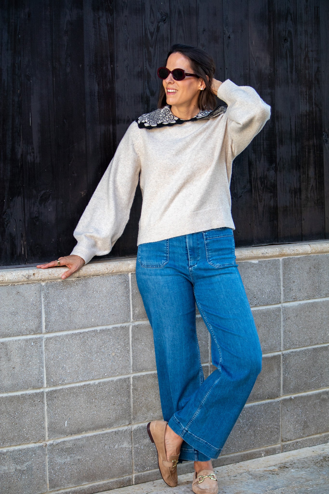 Woman wearing a tan sweater and blue jeans leaning against a wall.