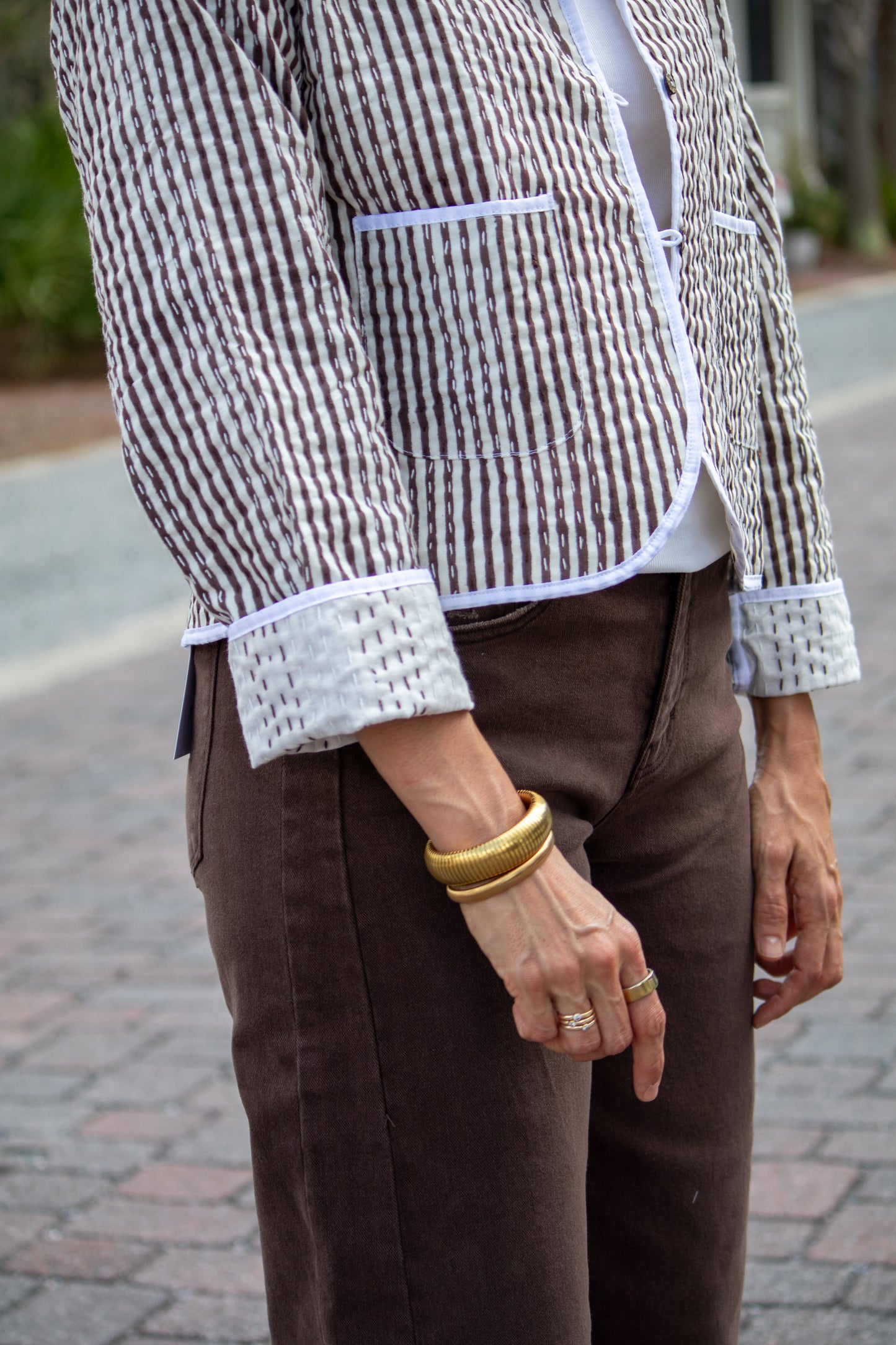Person wearing a patterned blazer, white shirt, brown pants, and gold bracelets on a blurred outdoor background.