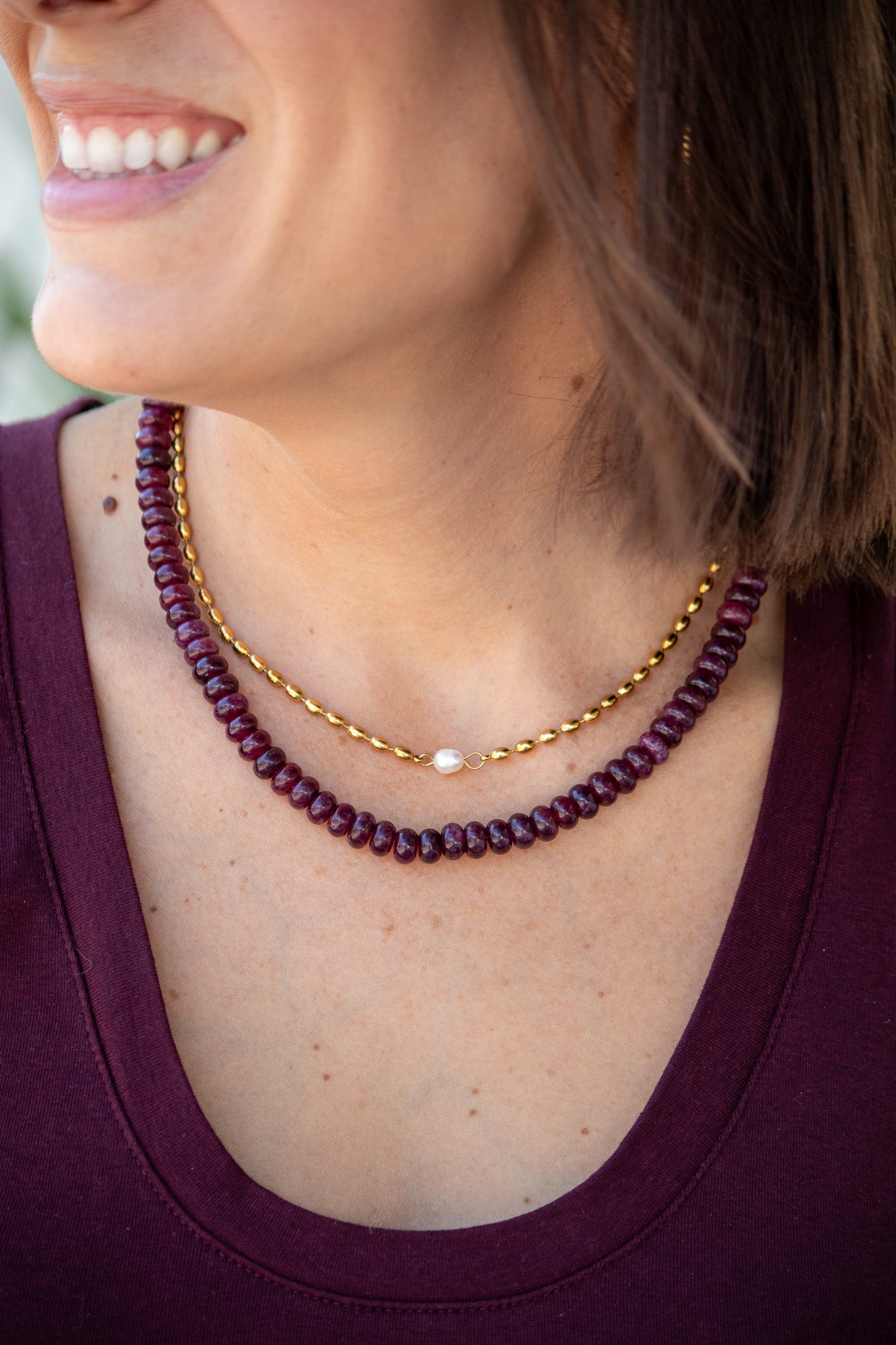 Close-up of a woman wearing a gold necklace and a berry color beaded necklace.