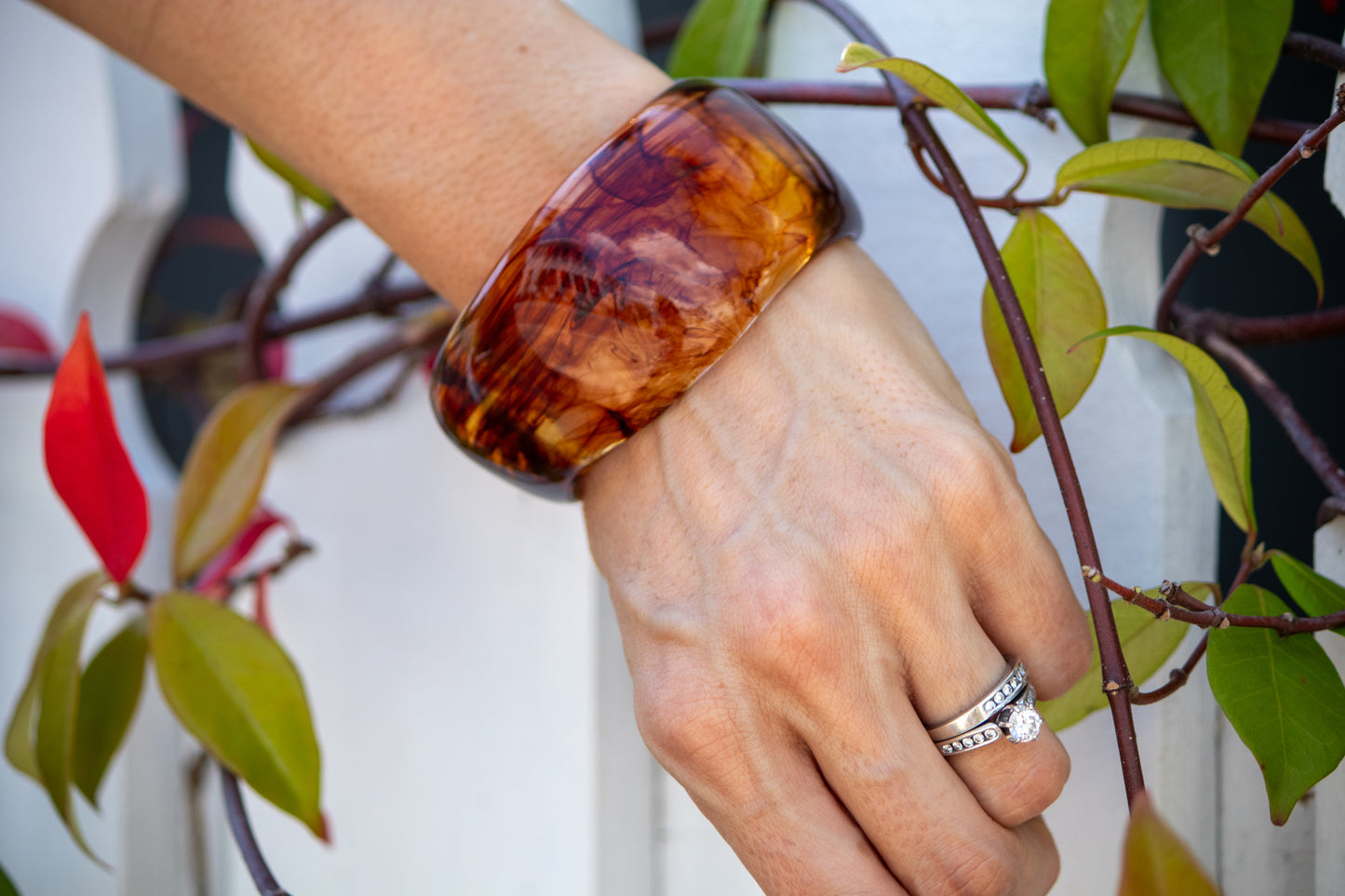 Hand wearing a tortoiseshell bracelet with a blurred background of green leaves and red flowers.