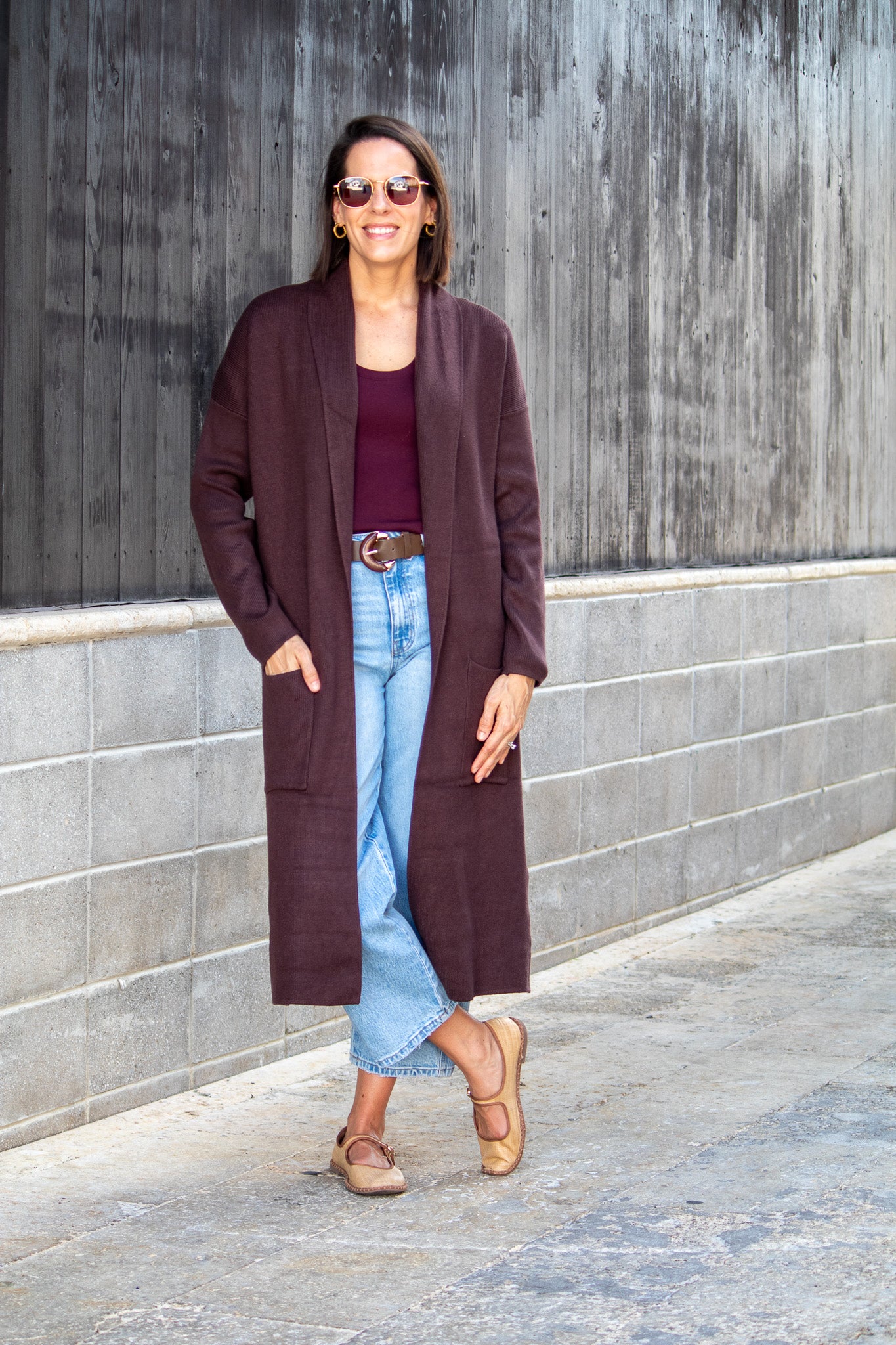 Woman wearing a long brown coat, maroon top, and light blue jeans against a gray wall.