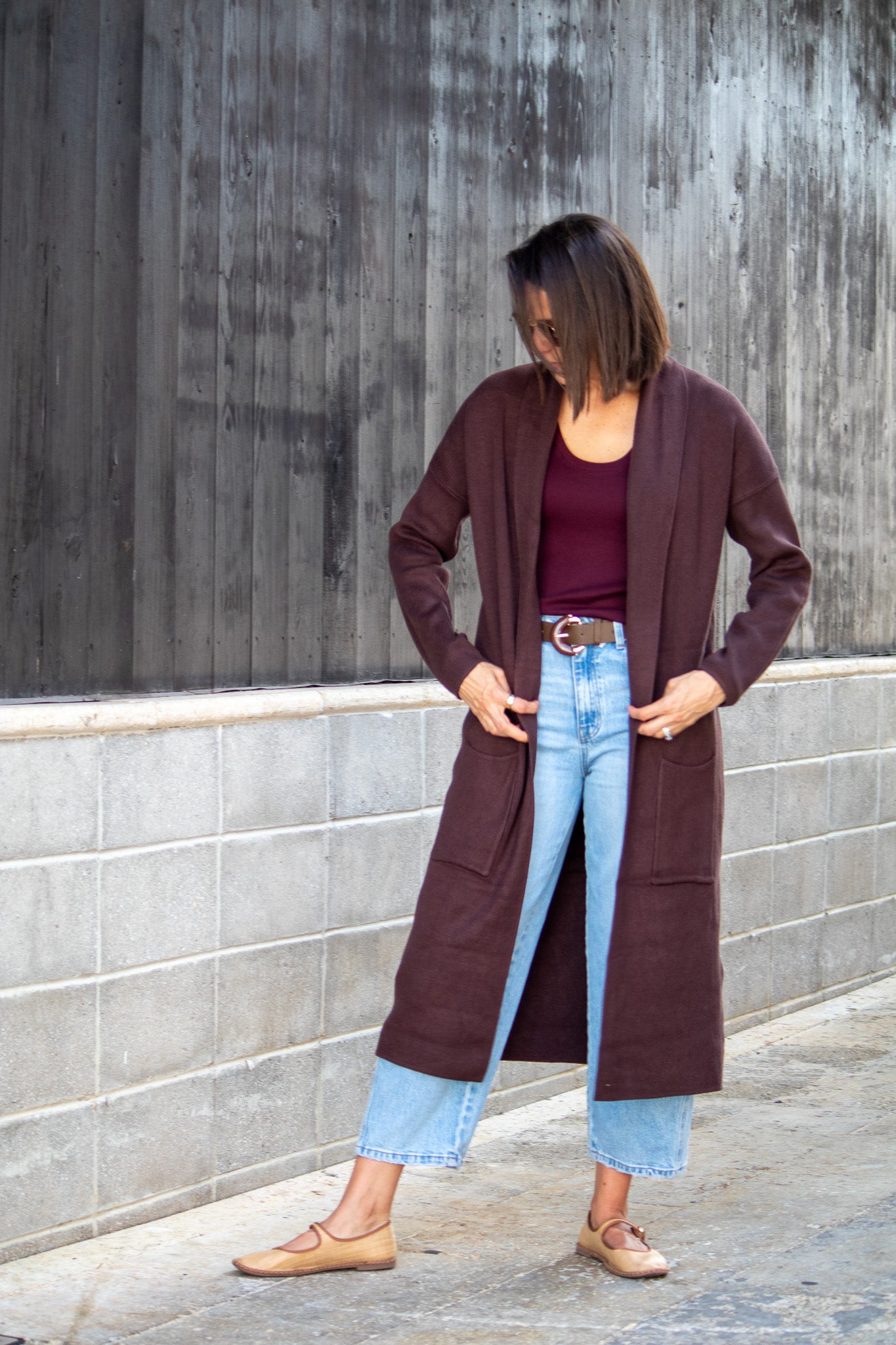 Woman wearing a long brown cardigan, burgundy top, and light blue jeans against a gray wall.