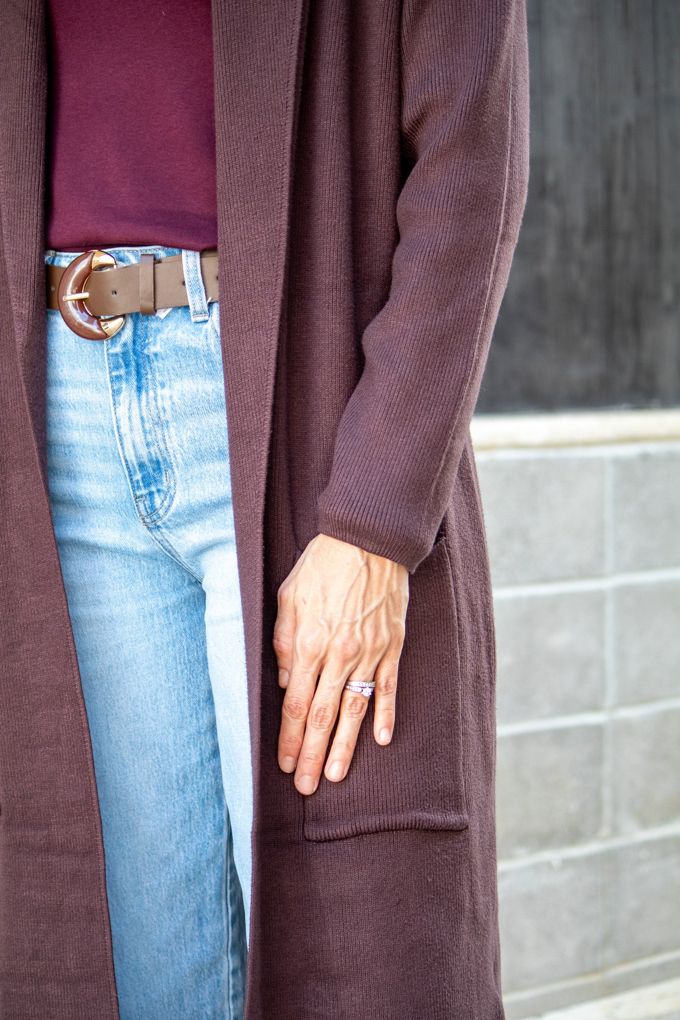 Person wearing a long brown cardigan, blue jeans, and a maroon shirt with a blurred background.