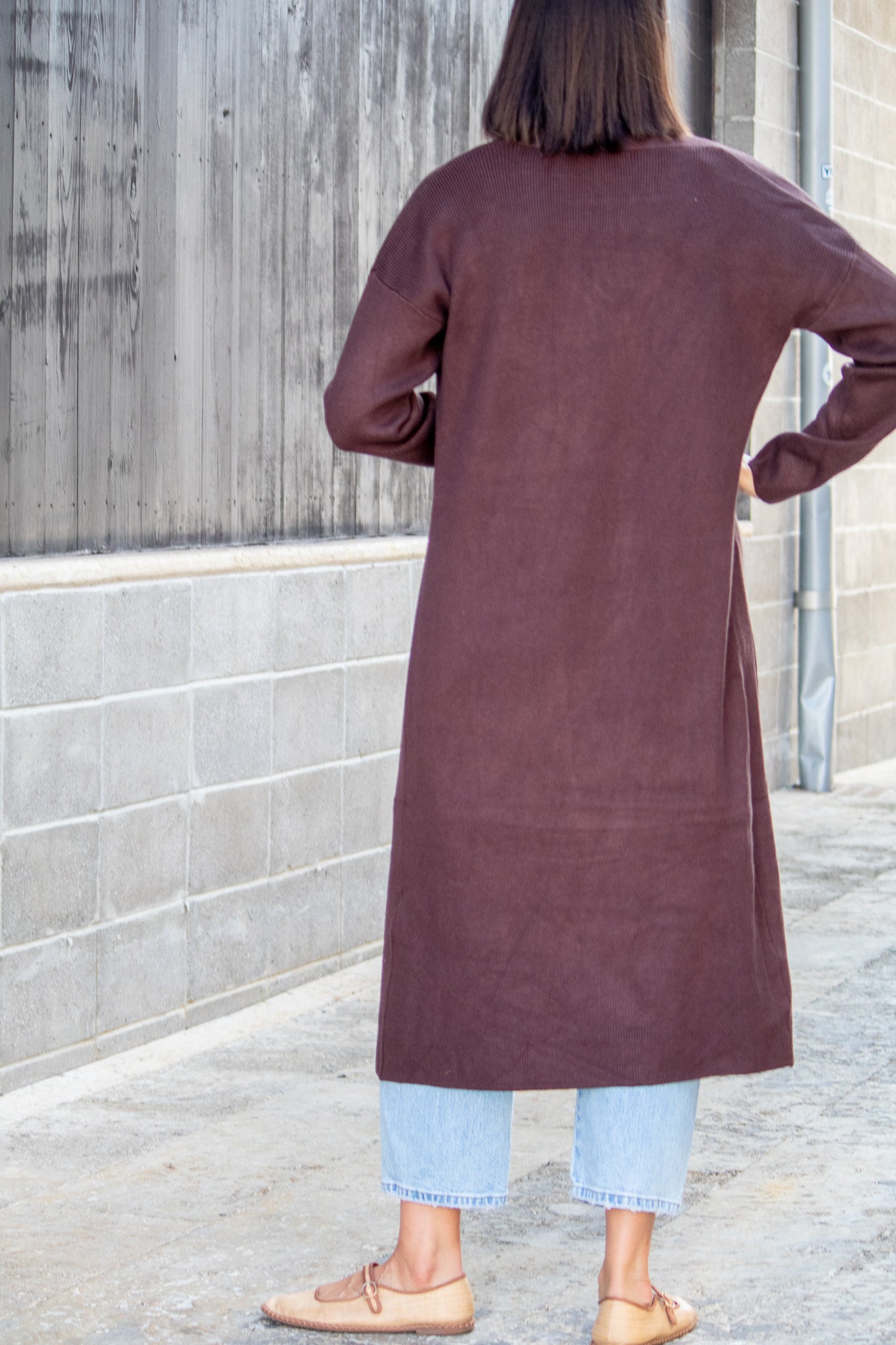 Person wearing a long brown coat standing against a gray wall.