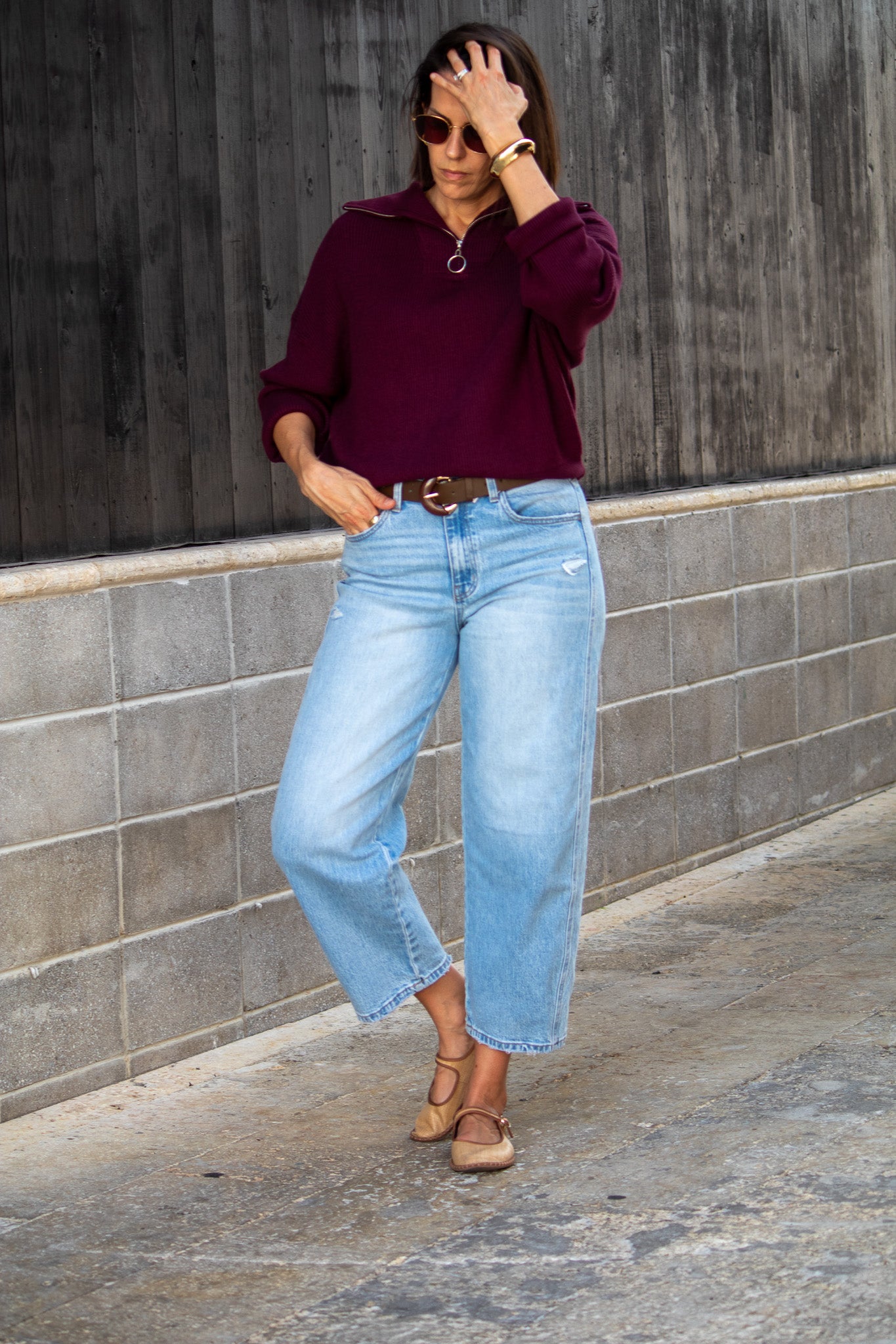 Woman wearing a burgundy sweater and light blue jeans against a gray wall.