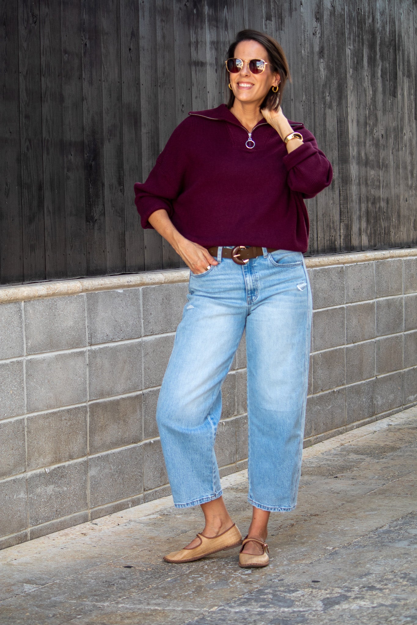 Woman wearing a purple sweater and light blue jeans standing against a gray wall.