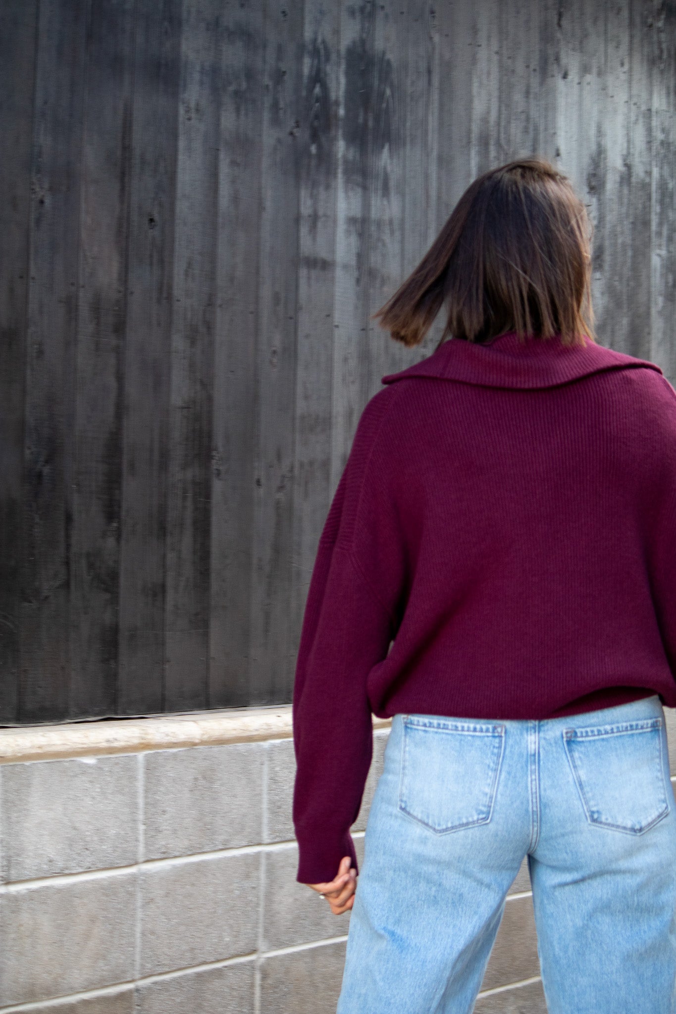 Person wearing a maroon sweater and light blue jeans standing against a concrete wall.