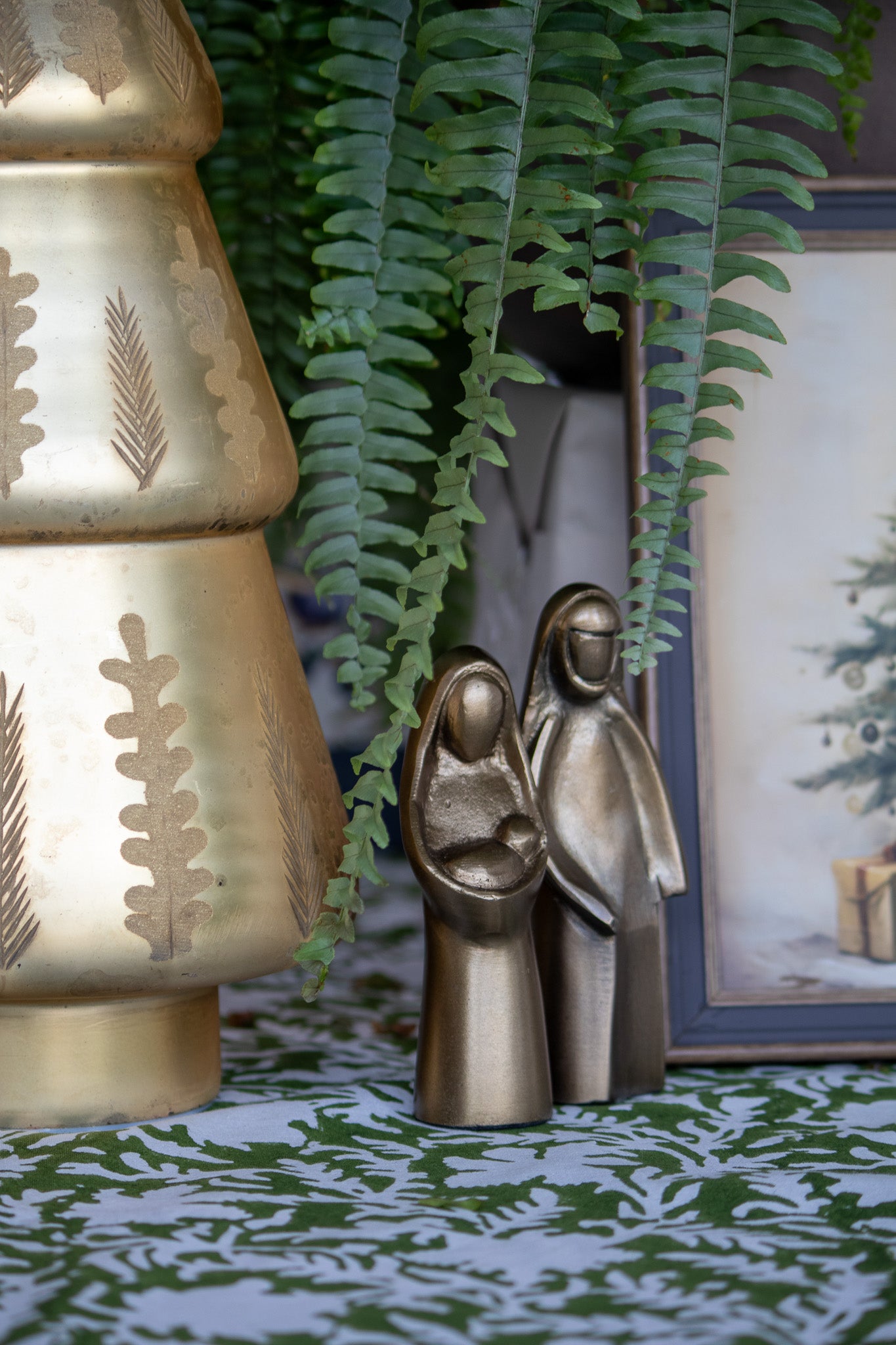 Bronze nativity scene figurines next to a gold Christmas tree with leaf patterns, against a green plant and framed picture background