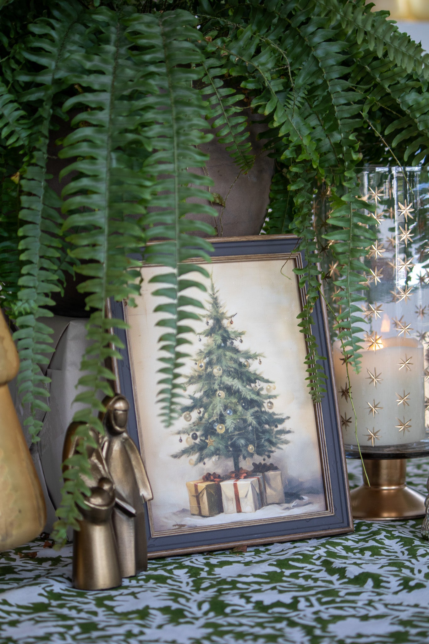 Ferns and a framed Christmas tree print with presents on a decorative table.