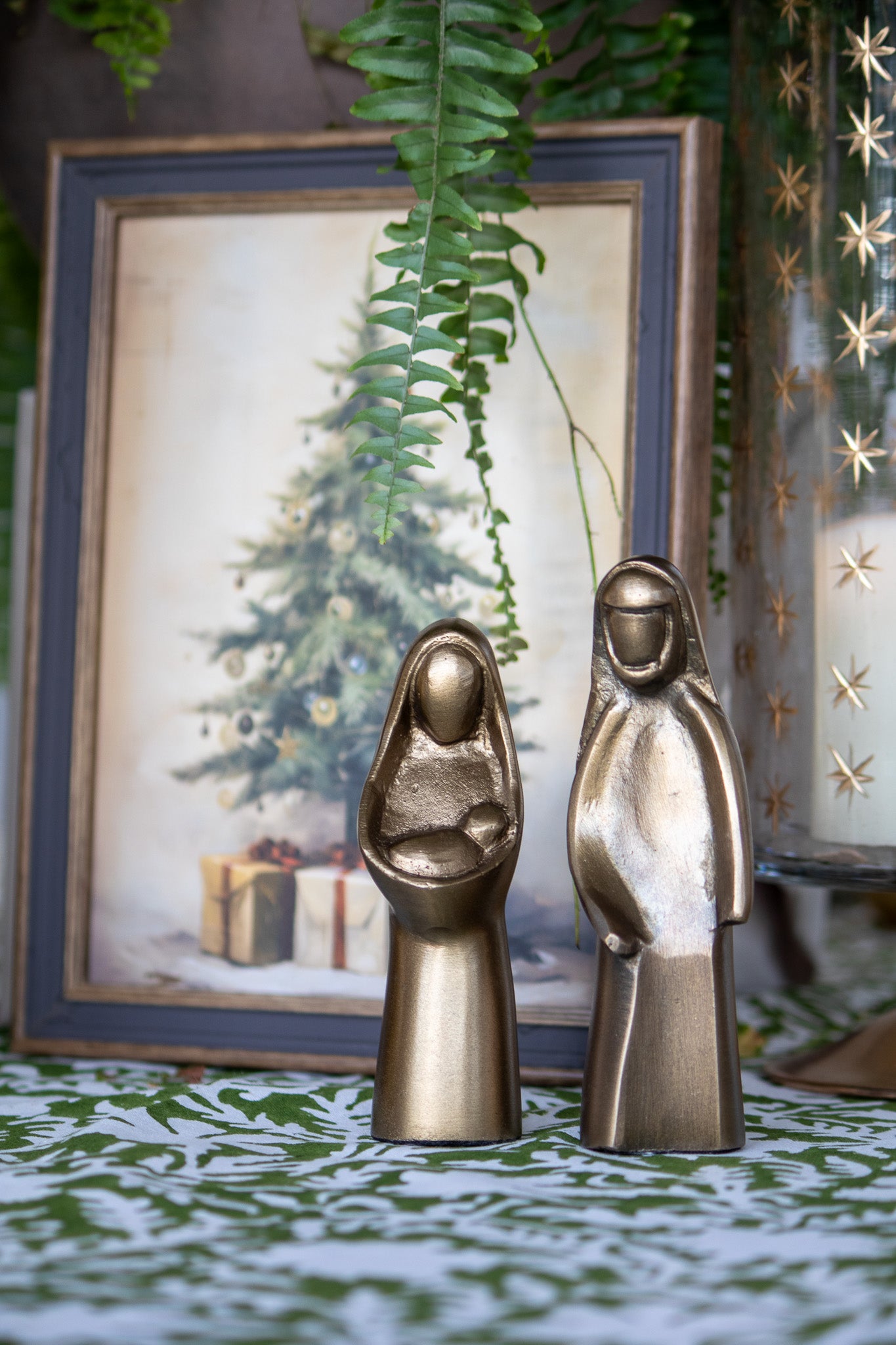 Bronze nativity scene figures with a framed Christmas scene in the background.