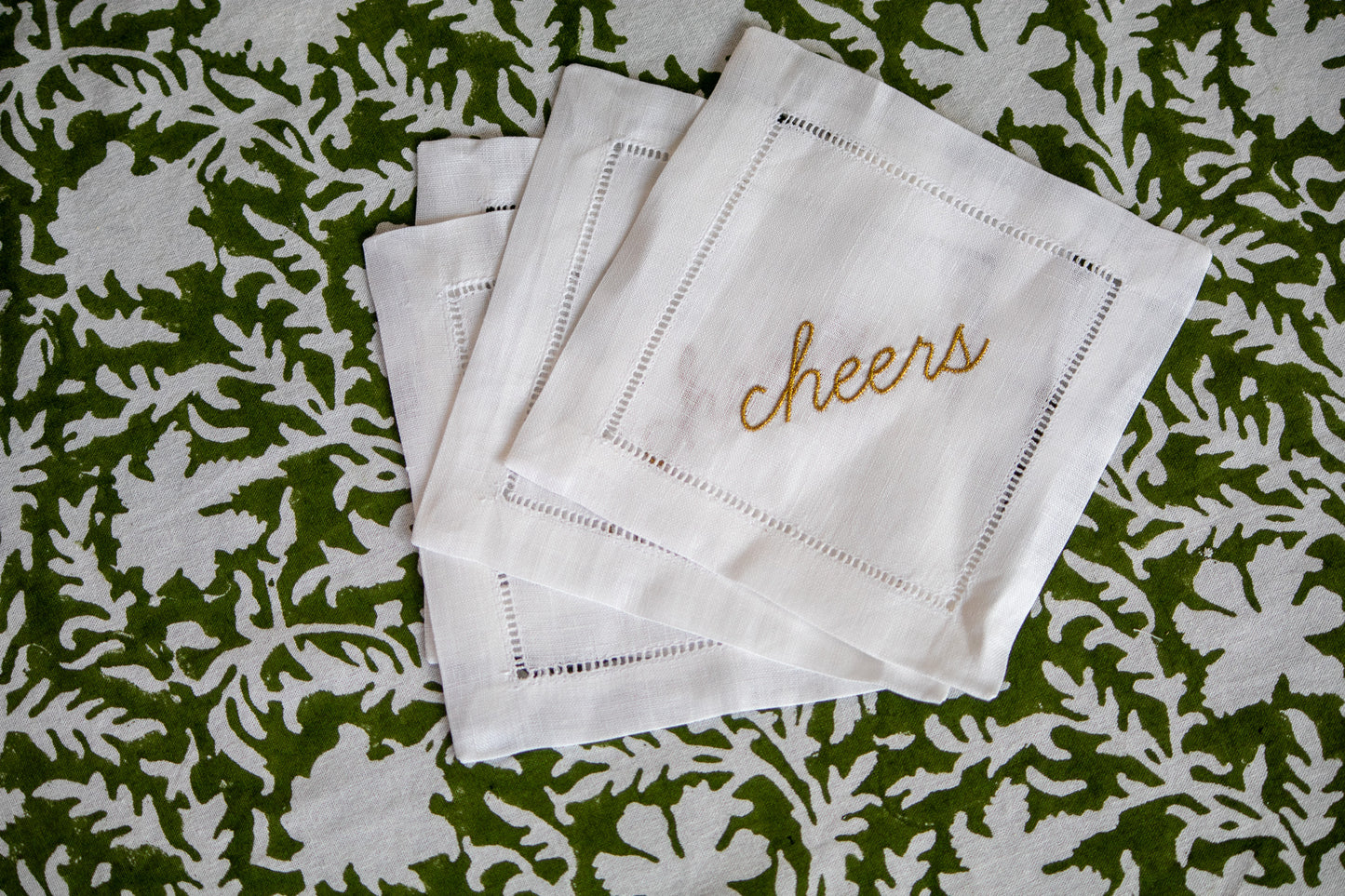 Set of white cocktail napkins with 'cheers' embroidery on a green floral patterned background