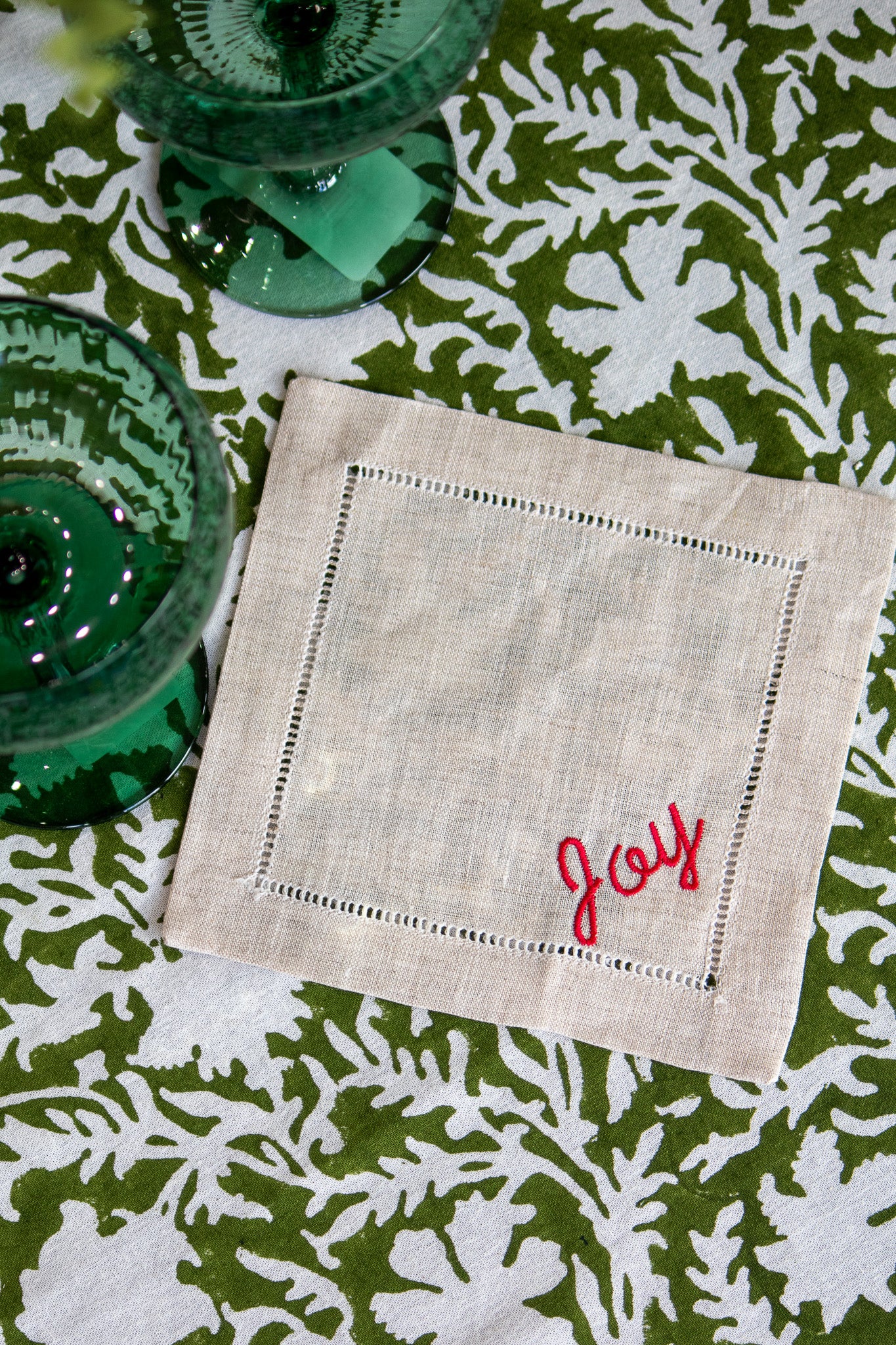 Linen coasters with 'Joy' embroidery on a green and white patterned tablecloth.