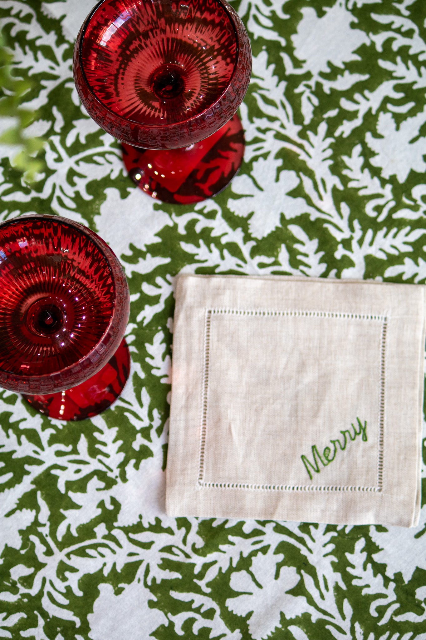 Two red glasses and a beige coaster with 'Merry' on a green and white patterned tablecloth.