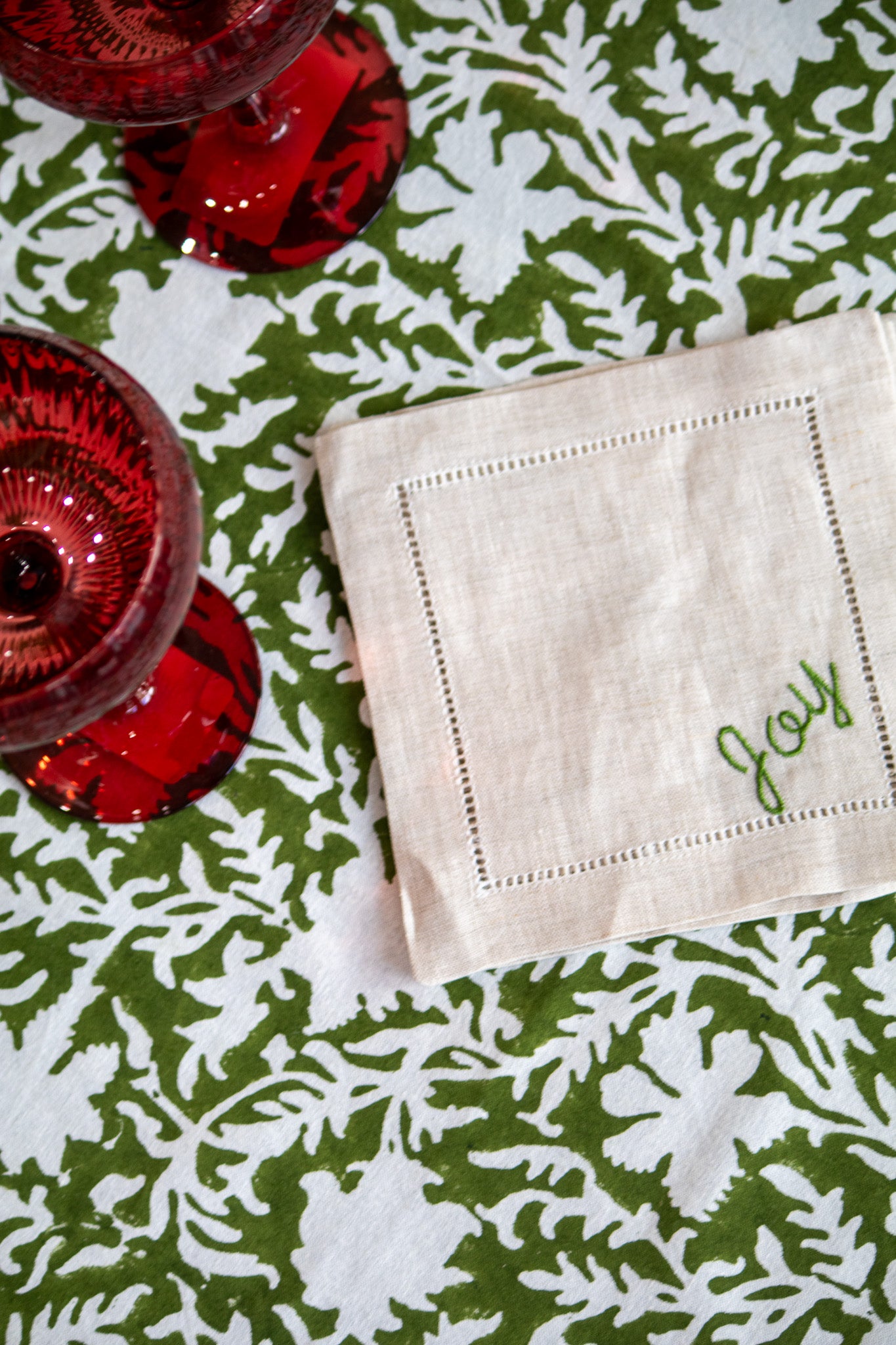 Coaster with 'Joy' embroidery on a green floral tablecloth with red glassware.