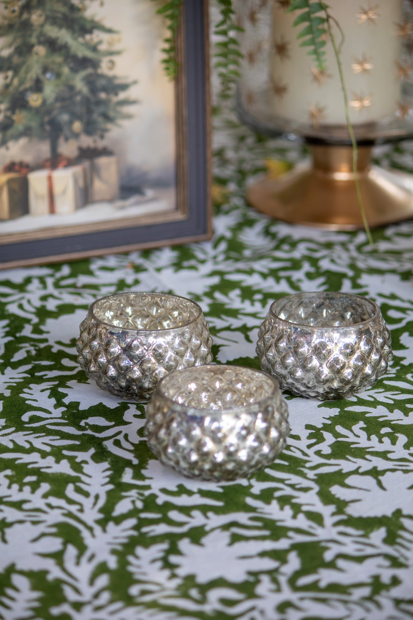 Three silver textured candle holders on a green and white patterned surface with a framed picture and candle in the background.