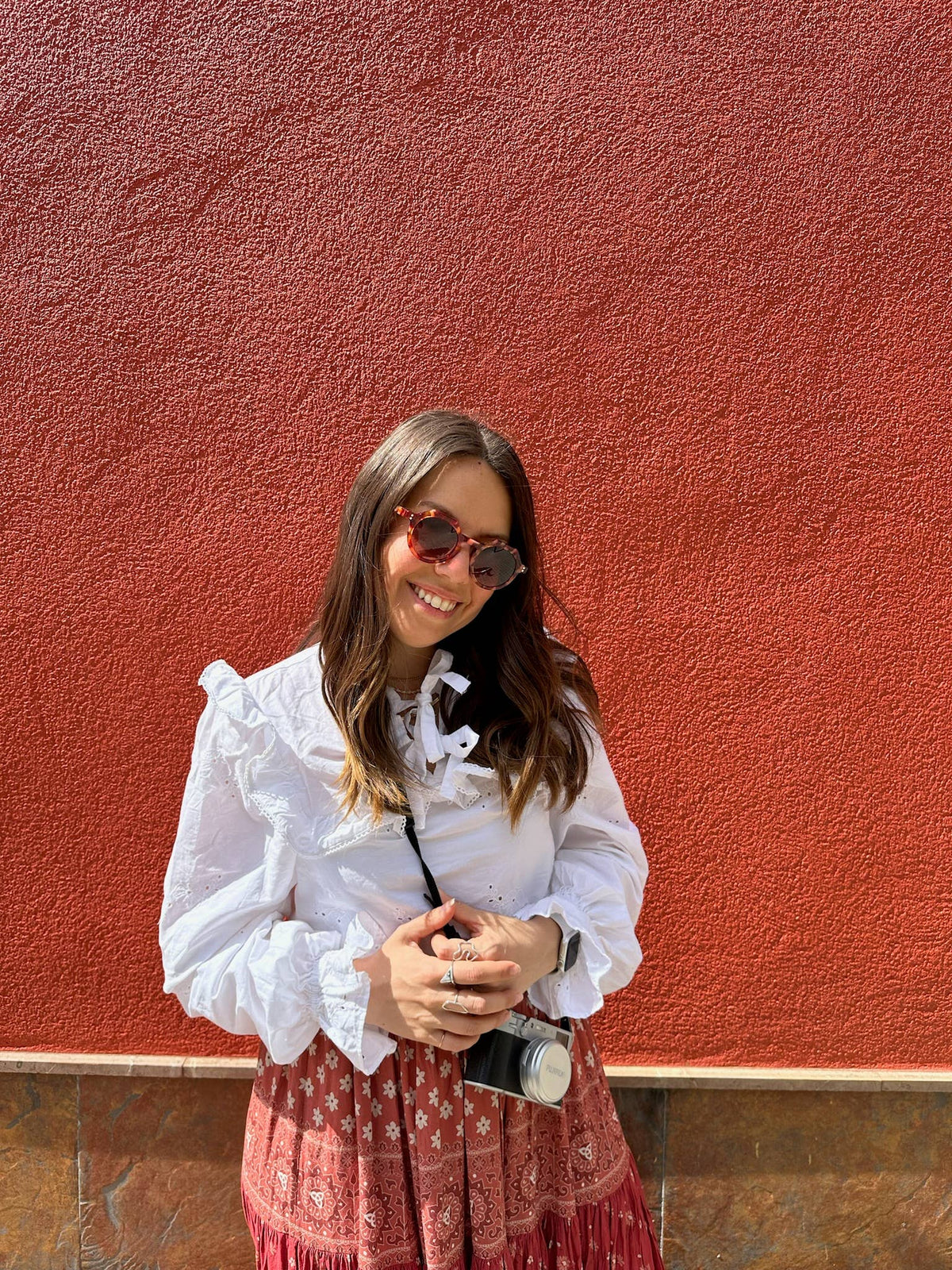 Woman in a white blouse and red patterned skirt standing against a red wall.