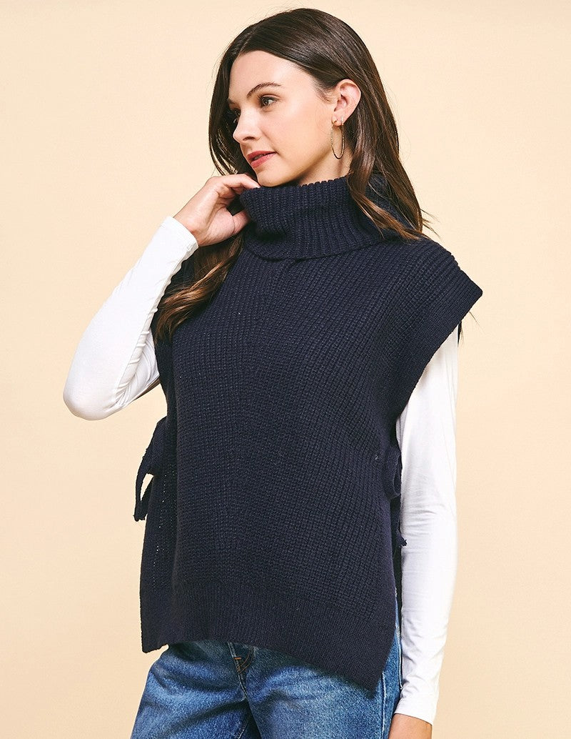 Woman wearing a navy blue knitted vest over a white long-sleeve shirt on a beige background