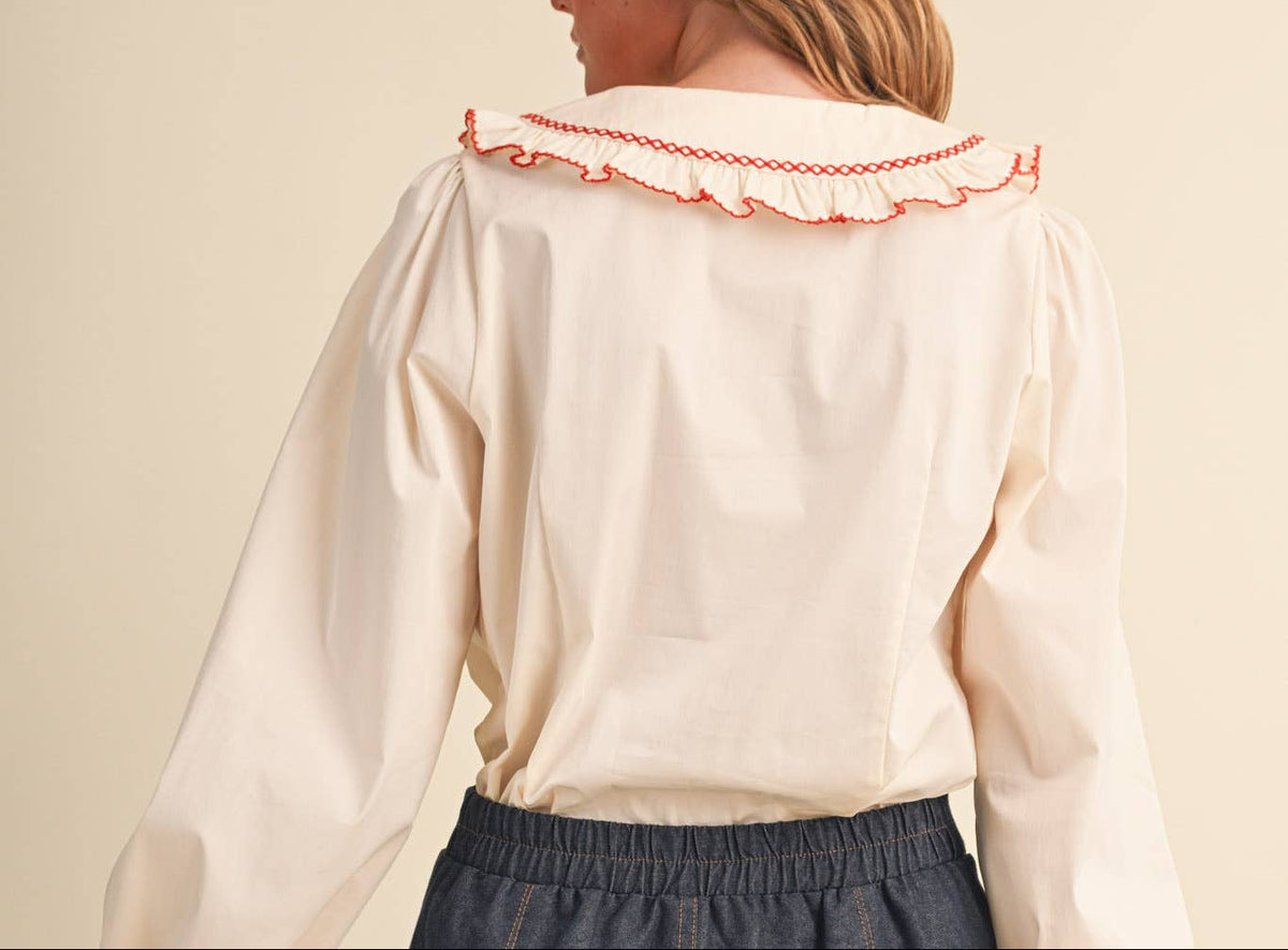 A woman shown from the back wearing a cream-colored blouse with a ruffled collar and cuffs trimmed in red, paired with a dark gray skirt, against a solid beige background.