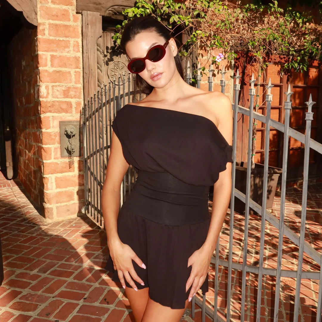 Woman wearing a black outfit with sunglasses standing in front of a brick wall and metal gate.
