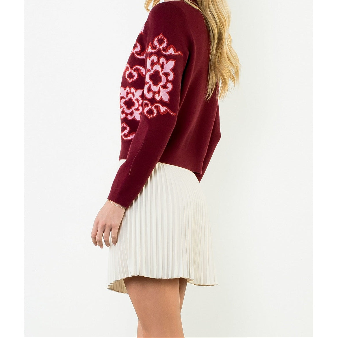 A woman modeling a burgundy knit sweater with a pink and white floral pattern on the sleeve, paired with a cream pleated mini skirt, against a plain white background.