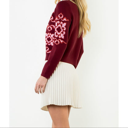 A woman modeling a burgundy knit sweater with a pink and white floral pattern on the sleeve, paired with a cream pleated mini skirt, against a plain white background.