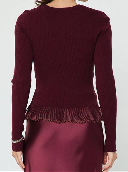 A woman shown from the back wearing a long-sleeve ribbed knit top in deep burgundy with a ruffled hem, paired with a matching satin skirt against a plain light gray studio background.