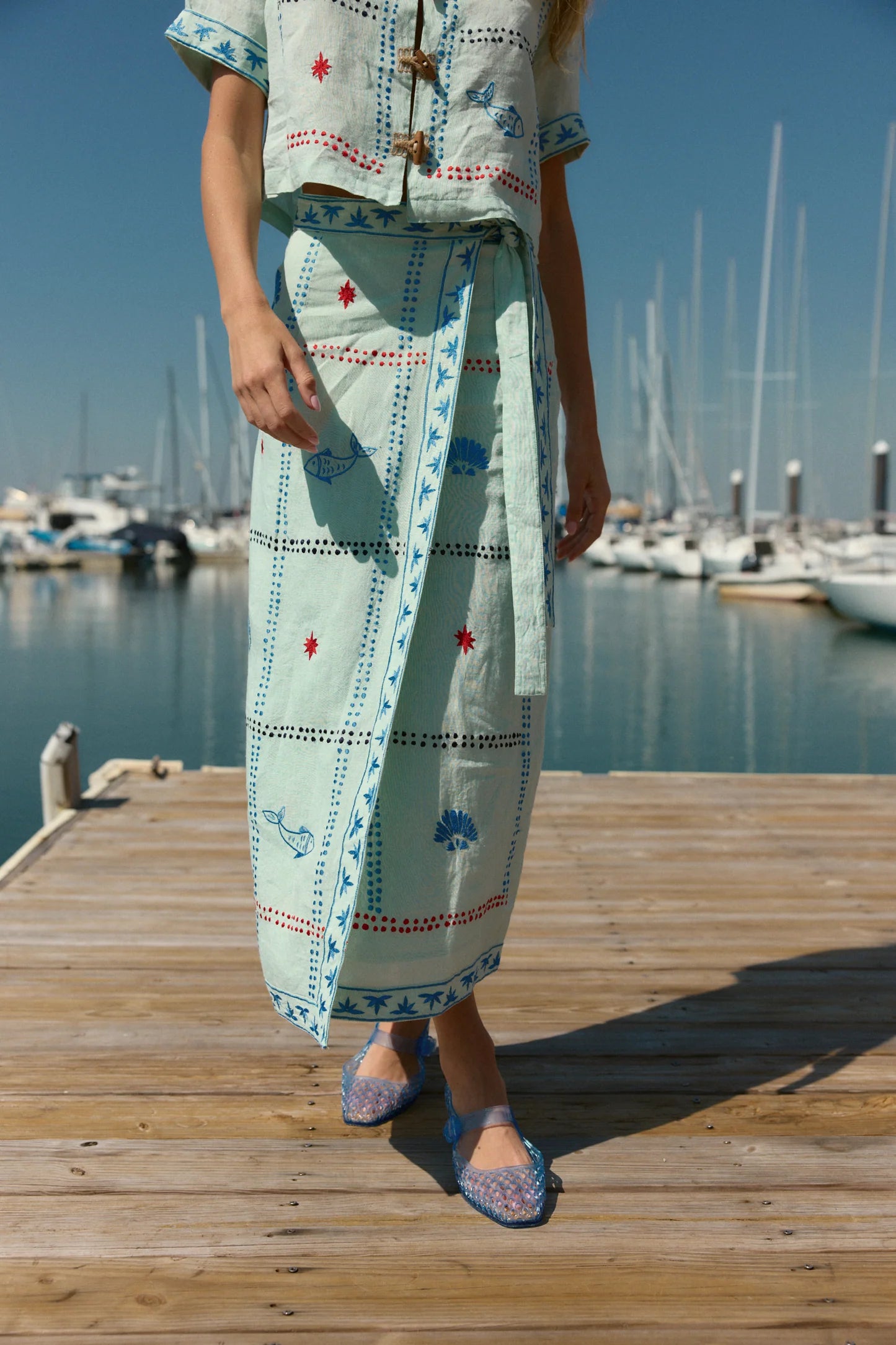 Person wearing a light blue embroidered outfit standing on a wooden dock with boats in the background.