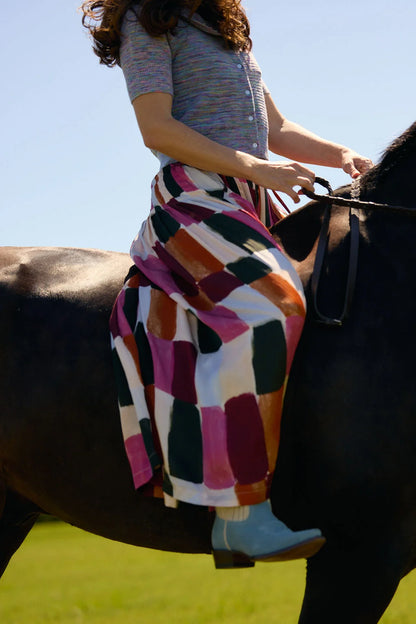A woman riding a dark horse outdoors, wearing a colorful geometric patterned pants with pink, orange, white, and dark green blocks, a gray knit top, and light blue boots.