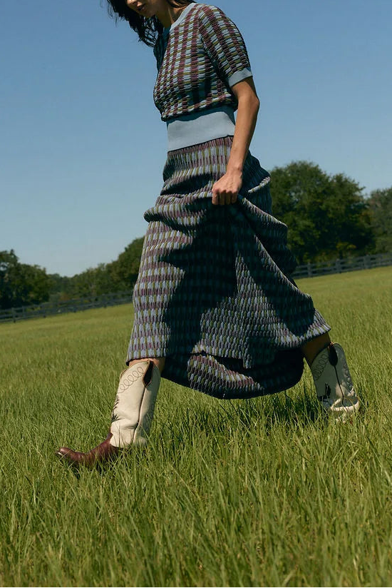 A woman walking across a green grassy field under a clear blue sky, wearing a dark patterned short-sleeve top and a long matching skirt with light-colored socks.