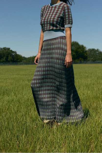 A woman stands in a grassy field under a clear blue sky, wearing a short-sleeve patterned top and a long pleated skirt with a geometric multicolor design.