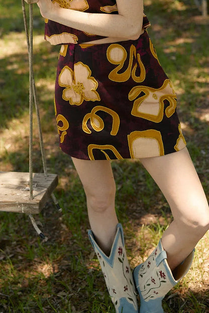 A woman stands outdoors in a park-like setting with trees, grass, and picnic tables, wearing a sleeveless dark skirt set with a bold yellow and tan abstract floral pattern and light blue western-style boots.