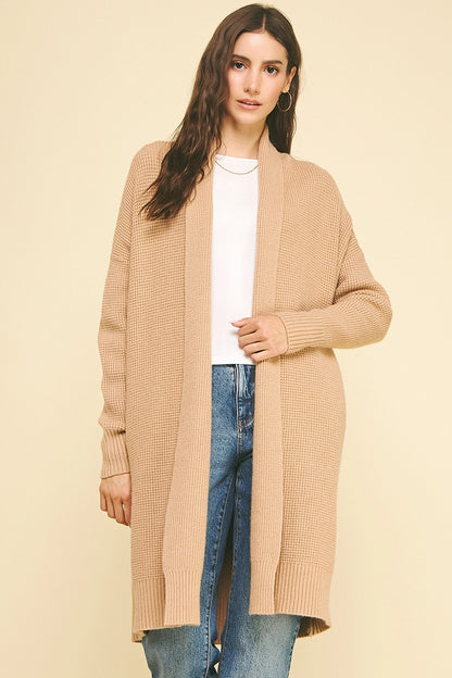 Woman wearing a long beige cardigan over a white top and blue jeans on a beige background