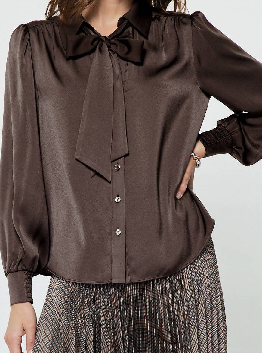 A woman wearing a dark brown satin blouse with a large bow at the collar and a button front, paired with a patterned skirt, against a light gray studio background.